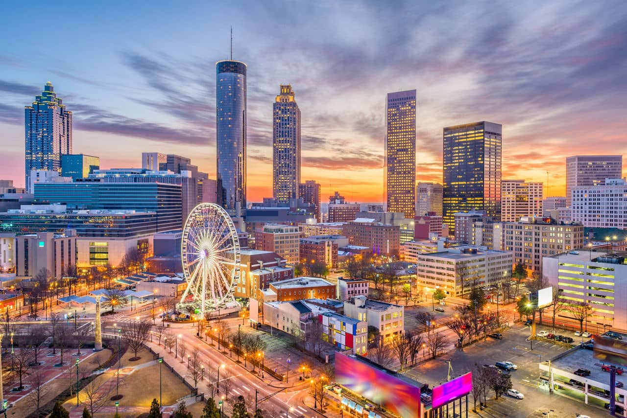 Downtown Atlanta, GA skyline at sunset with lit skyscrapers, Ferris wheel, and Centennial Park under colorful evening sky.
